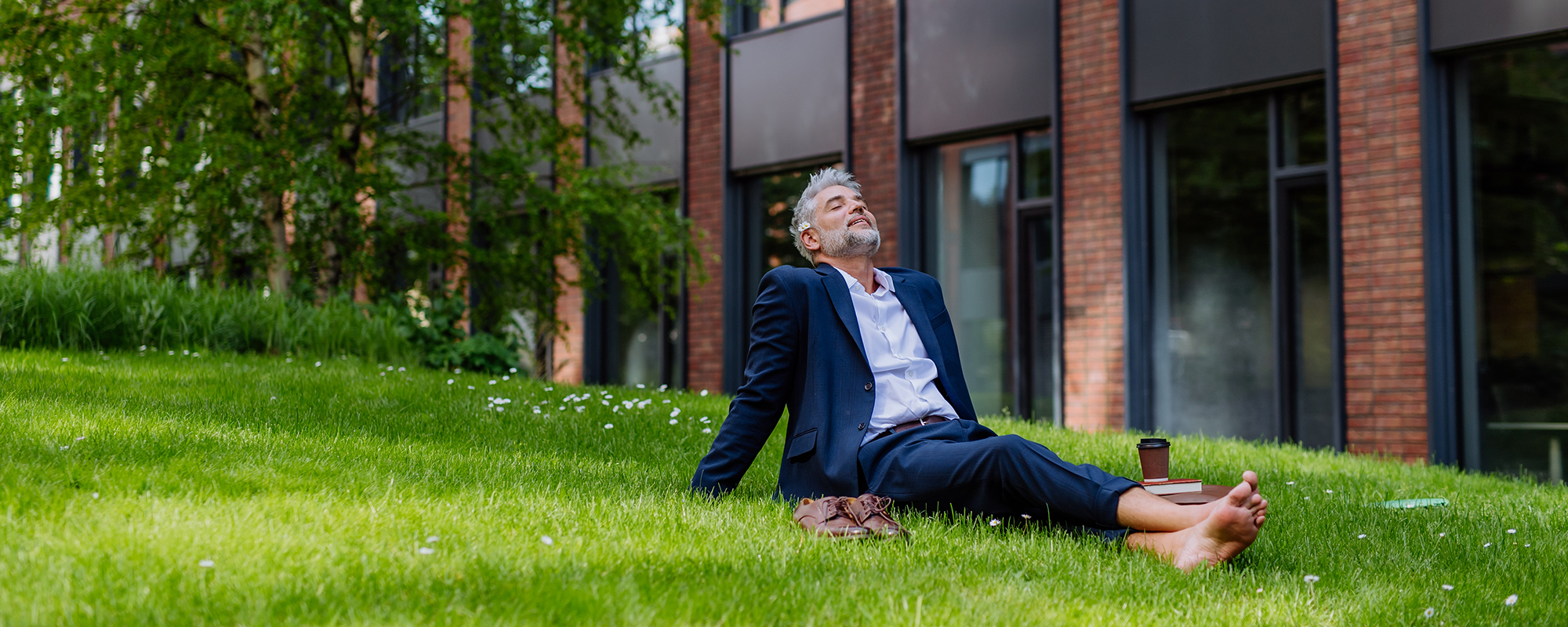 attraktiver Mann mittleren Alters in Businessanzug und barfuß sitzt auf grüner Wiese vor Bürogebäude und entspannt sich