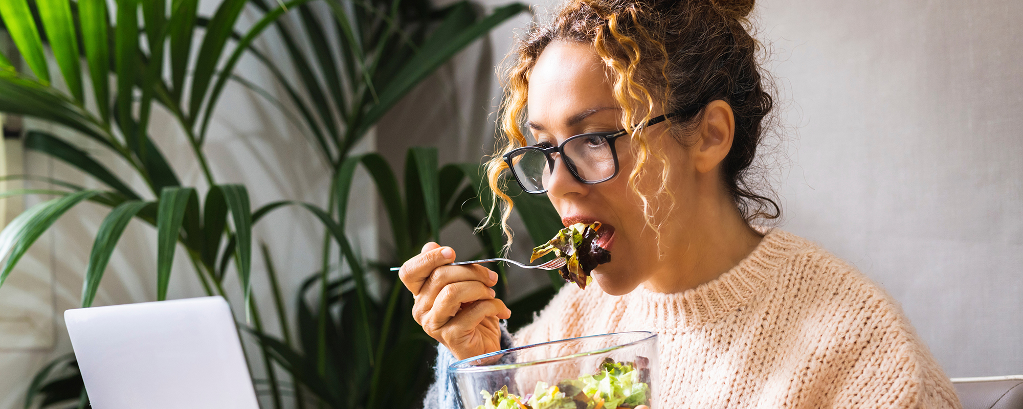 junge Frau mit blonden, hochgesteckten Locken und schwarzer Hornbrille sitzt vor PC und isst Salat aus einer Glasschüssel