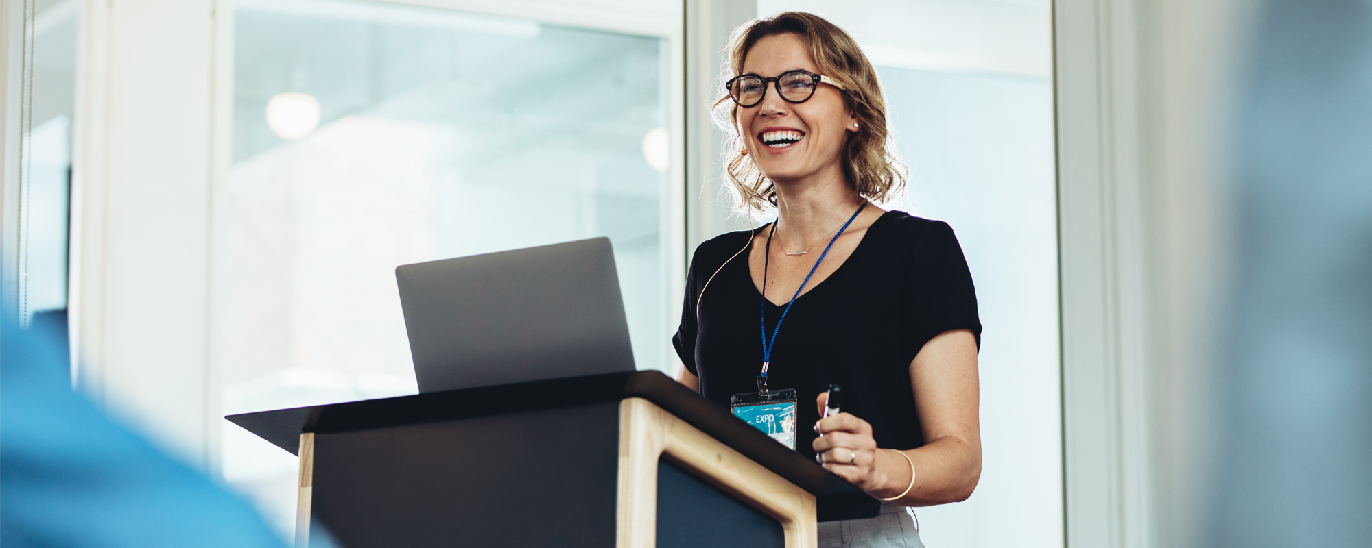 junge blonde Frau mit Brille steht lachend an Rednerpult, aufgeklappter Laptop für Präsentation steht am Pult
