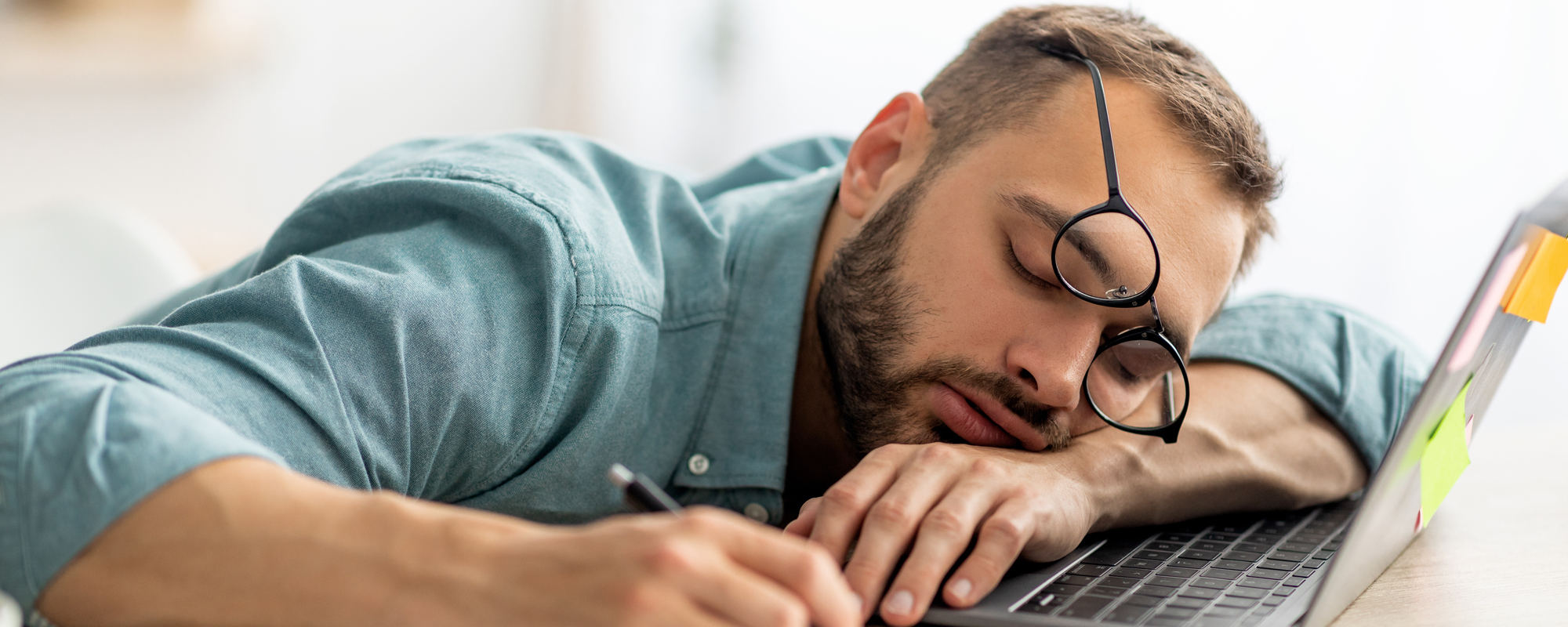 Schon wieder Überstunden. Junger braunhaariger Mann mit Vollbart schläft auf Laptop-Tastatur ein. Brille verschoben, Kuli noch in der Hand.