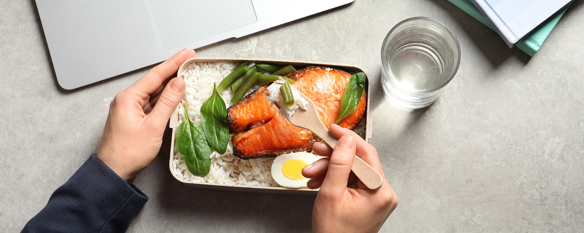 Person am einem Arbeitstisch isst ein gesundes Mittagessen aus Reis, Lachs und Gemüse aus einer Lunchbox.