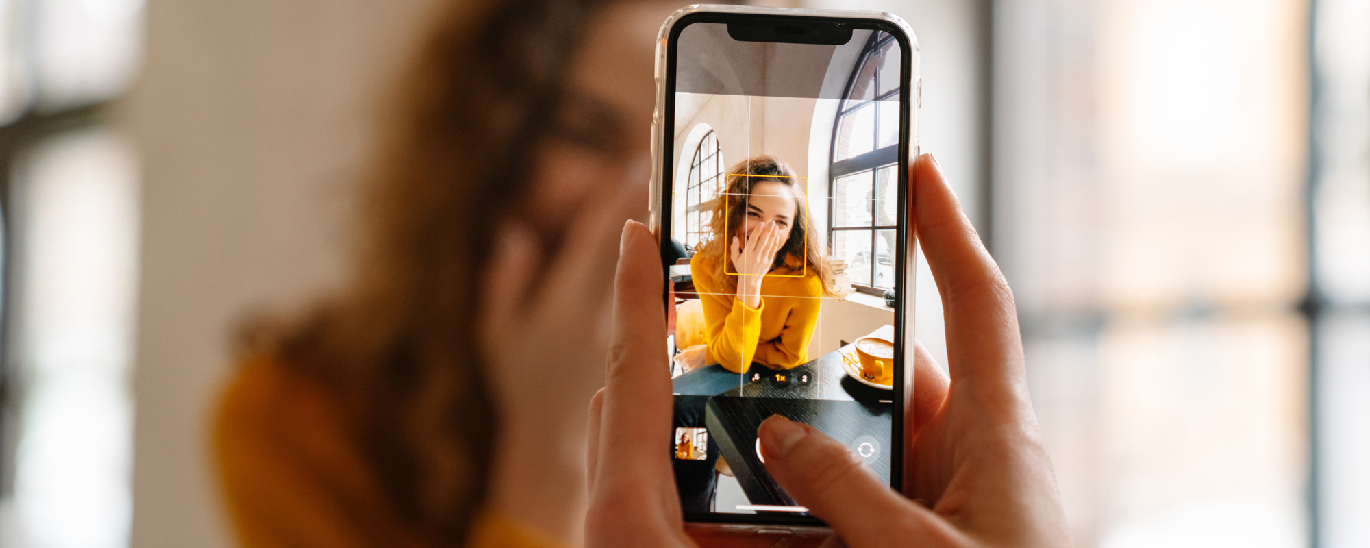 Gutes Bewerbungsfoto? Frau in gelbem Oberteil lacht mit Hand vor dem Mund, während sie mit einem Handy fotografiert wird.